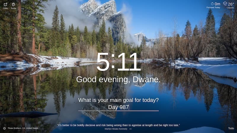 A wide shot of a lake reflecting the snow-capped peaks of the Sierra Nevada mountain range. The water is calm and still, and the snow on the ground reflects the light of the rising sun. The image is taken from the edge of a forest, looking towards the mountains, a trail winds along the edge of the lake. The location is "Three Brothers, CA, United States".
