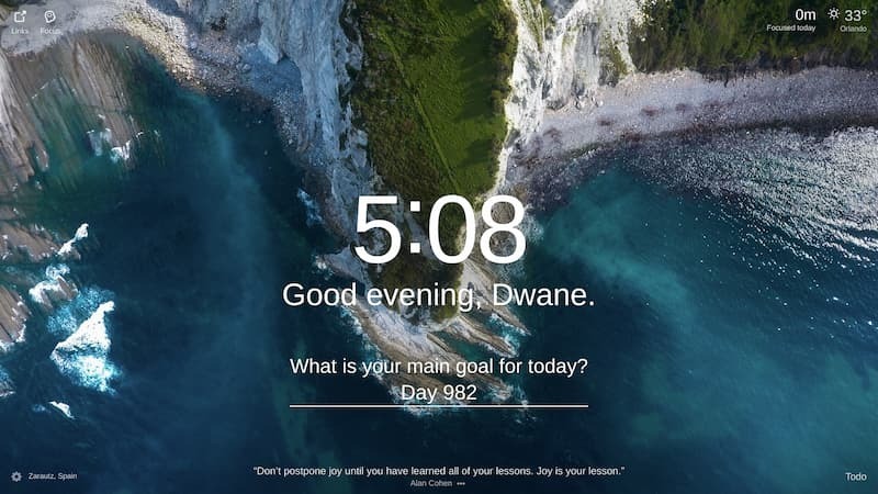 An aerial view of a rocky shoreline, with turquoise ocean water lapping at the shore. The rocks are white and jagged, and there is a small patch of green grass at the top of the cliff. The image is taken from a high angle, so the ocean appears vast. Location: Zarautz, Spain