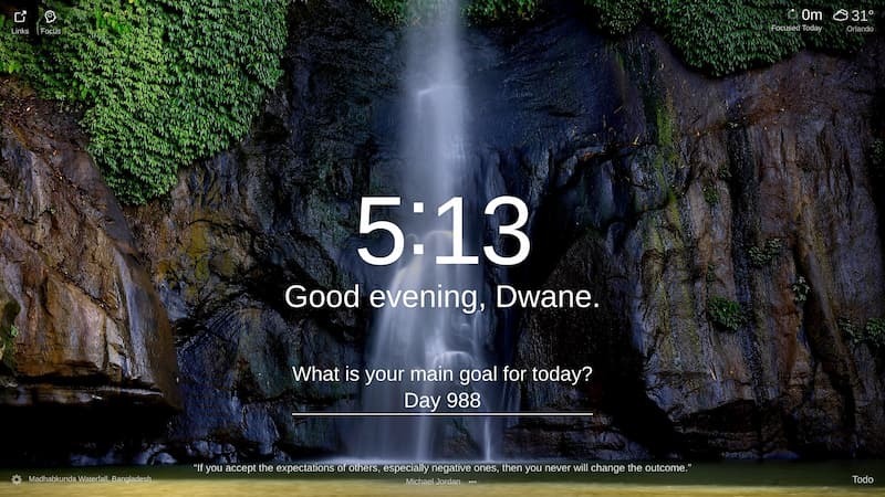 A large waterfall cascades down a rock face, with the mist from the water obscuring part of the cliff face. Vines are visible on the left and right of the waterfall. The rock face is dark and rugged. Location: Madhabkunda Waterfall, Bangladesh.