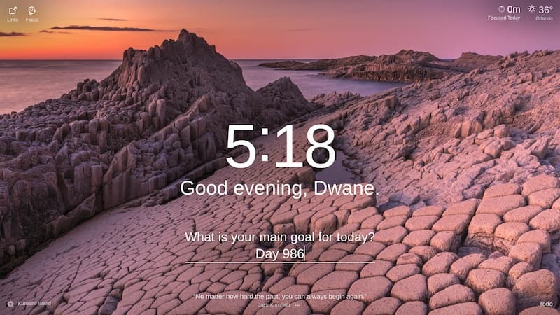 A photo of a rocky, coastal landscape with a body of water in the background, a sunrise or sunset is happening over the water, the landscape is a dark grey-purple color and the sky is a gradient of purples and oranges. The location is called Kunashir Island.