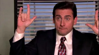The image depicts Michael Scott, a character from the TV show "The Office." He is in an office setting, wearing a suit and tie. Michael is making a playful gesture with both hands, holding up his pinky and index fingers while the rest of his fingers are curled down, mimicking a rock and roll sign. His facial expression is slightly serious yet amused, and the office blinds and window are visible in the background.