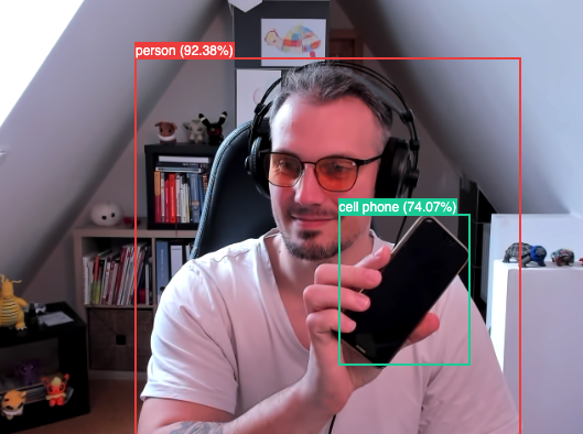 Detected objects are person and cell phone in a screenshot of a person sitting in a chair holding up a cell phone.