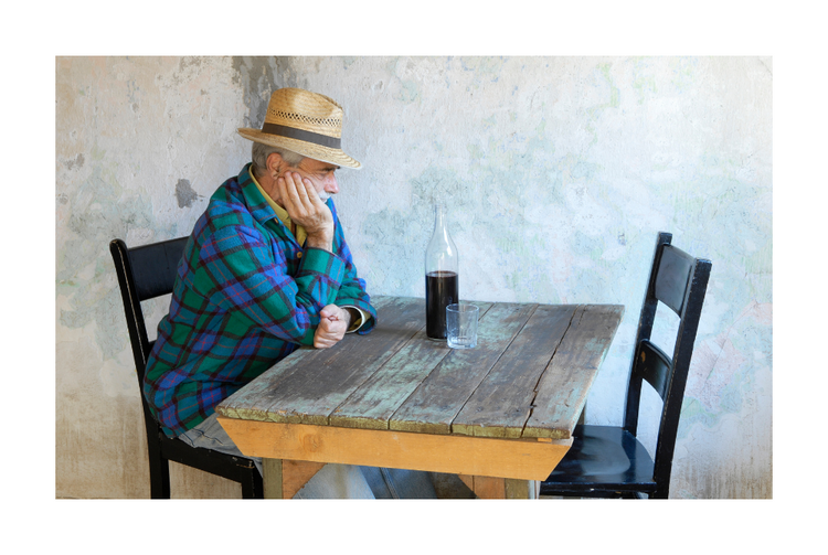 An older man is sitting alone on a table drinking some kind of liquid.