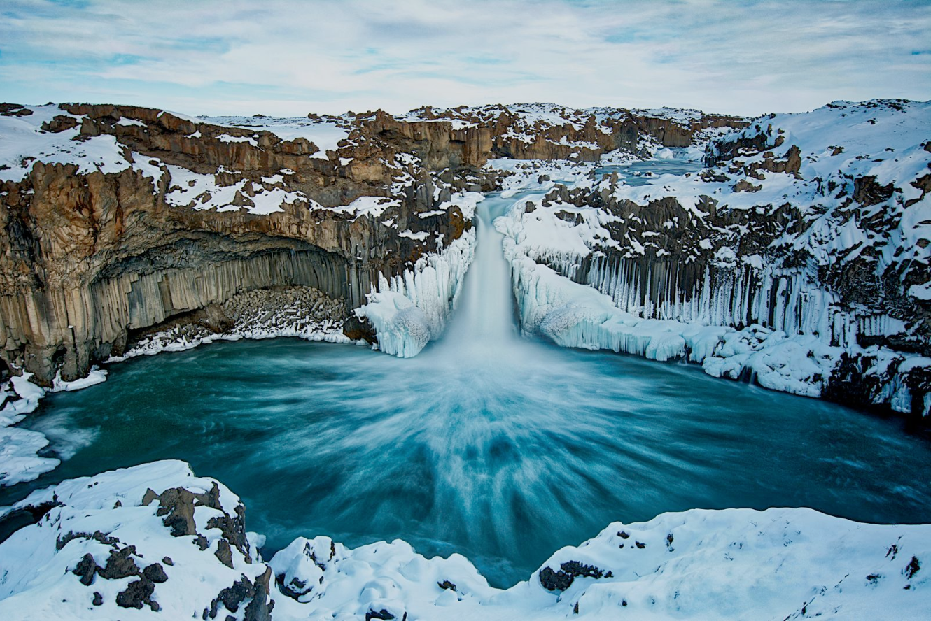 A photograph of a partially frozen fall with its pool in deep blue