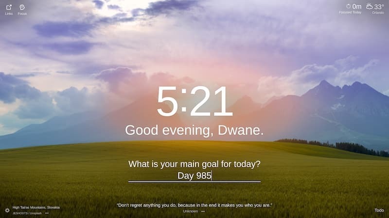 A panoramic shot of a rolling, green grassy field with a mountain range in the background. The sky is a vibrant blue with a hazy glow and streaks of pink and orange clouds. The scene is set at dusk, with a hazy orange glow over the mountains. The location is High Tatra Mountains, Slovakia.