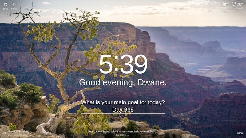 The image shows a rugged canyon landscape with a large, winding river running through it. The canyon walls are sheer. The sky is a light blue and there are some clouds in the distance. There is a tree in the foreground on a cliff edge. The location is the Grand Canyon in the United States.