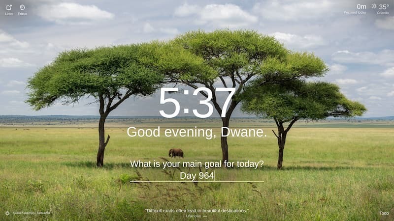 The image shows a vast, grassy plain in the Serengeti National Park in Silale Swamps, Tanzania. There are three large, green trees in the foreground, and a herd of elephants can be seen grazing in the distance. The sky is a clear blue, and the sun is shining brightly.