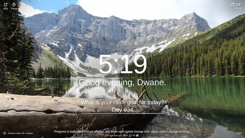 The photo shows a beautiful mountain lake in Banff National Park, Rawson Lake, AB, Canada. The water is a deep blue-green color and the sky is clear with a few clouds. The mountains in the background are covered in snow. There are trees on the shore of the lake and a large log juts out into the water on the left side of the photo.